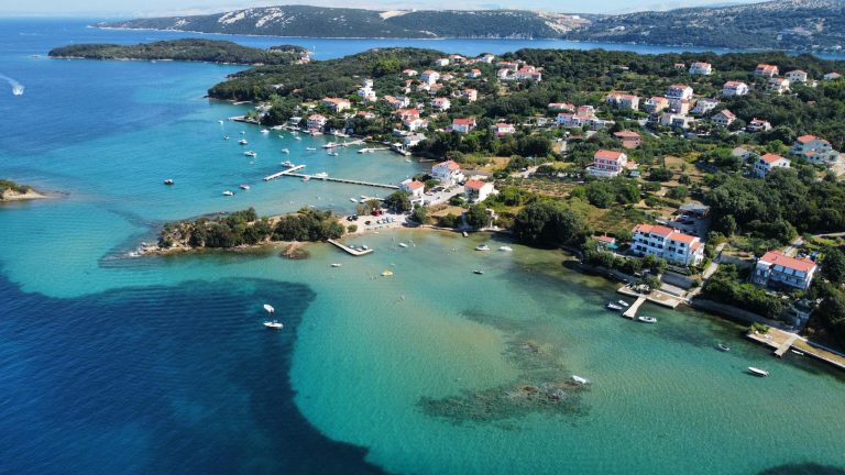 Explore the stunning coastline of Rab Island, Croatia, with turquoise waters and charming buildings from above.