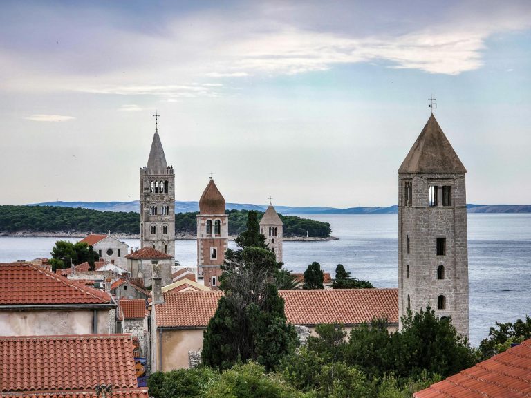 Scenic view of historic bell towers in Rab, Croatia against the serene Adriatic Sea and lush greenery.